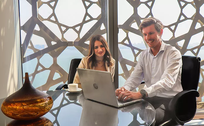 Two professionals smiling and working in a serviced office with laptops, overlooking a city view.